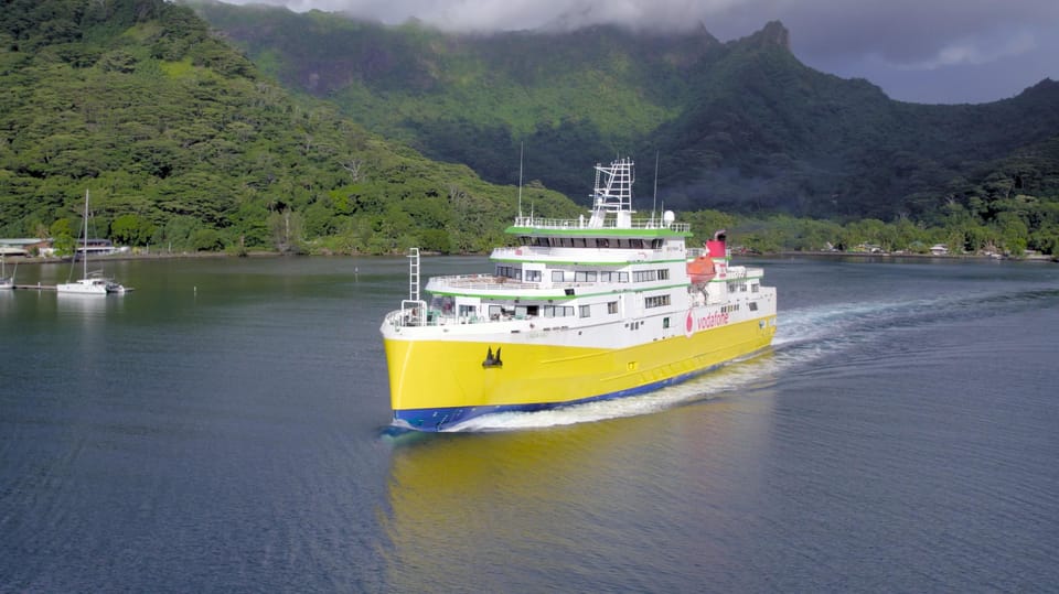 A white passenger ferry with an open upper deck crossing calm water, the green ridgeline of an island low on the horizon behind it