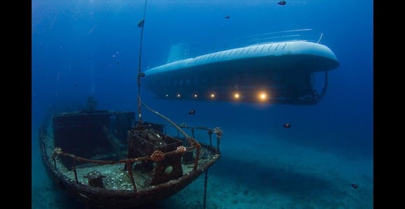 A white submarine hovering in deep blue water above the wreck of a small wooden sailing boat on a pale sandy seabed, its row of portholes lit from inside