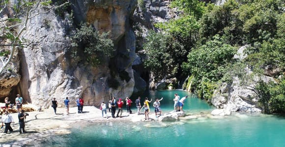 Turquoise canyon pool below grey limestone cliffs with visitors standing on a pale gravel bank among green trees