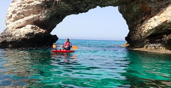 A kayaker in a red life jacket paddling a red kayak beneath a natural rock arch in clear green water