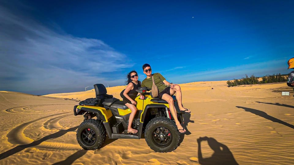 Quad jaune sur le sable des Dunes blanches, un couple posant dessus, une crête de dune, des buissons verts et un ciel bleu en arrière-plan