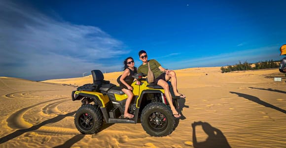 Yellow quad bike in the sand of the White Dunes, a couple posing on it, a dune ridge, green scrub and blue sky behind