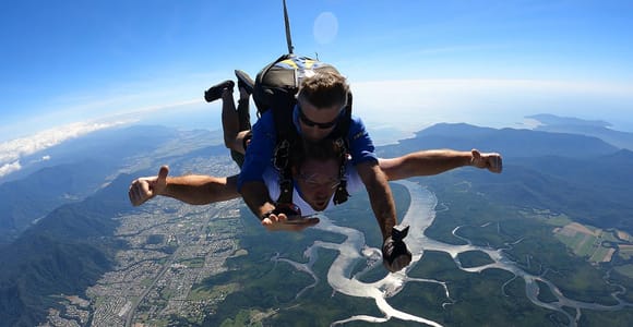 Two people in freefall high above the coast, the tandem instructor in a blue shirt with his arms spread wide, the town and green hills far below and a river winding in silver loops across a flat green plain