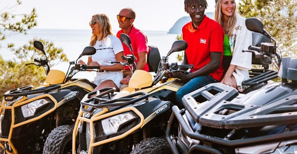 Four riders on yellow and black quad bikes stopped on a stony track, pine trees and the sea behind them