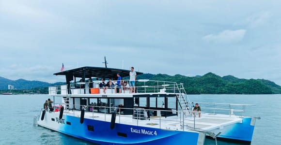A blue catamaran named Eagle Magic 4 at anchor in flat water, a handful of passengers on the shaded upper deck, Langkawi's forested hills behind under a grey sky