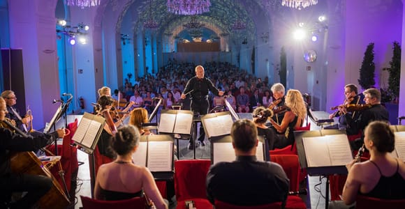 An orchestra and conductor playing in a long vaulted hall under chandeliers, with the audience seated in rows behind