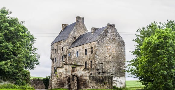 A weathered stone tower house with a slate roof and an arched gateway at the end of a country track, surrounded by grass and trees under a grey sky