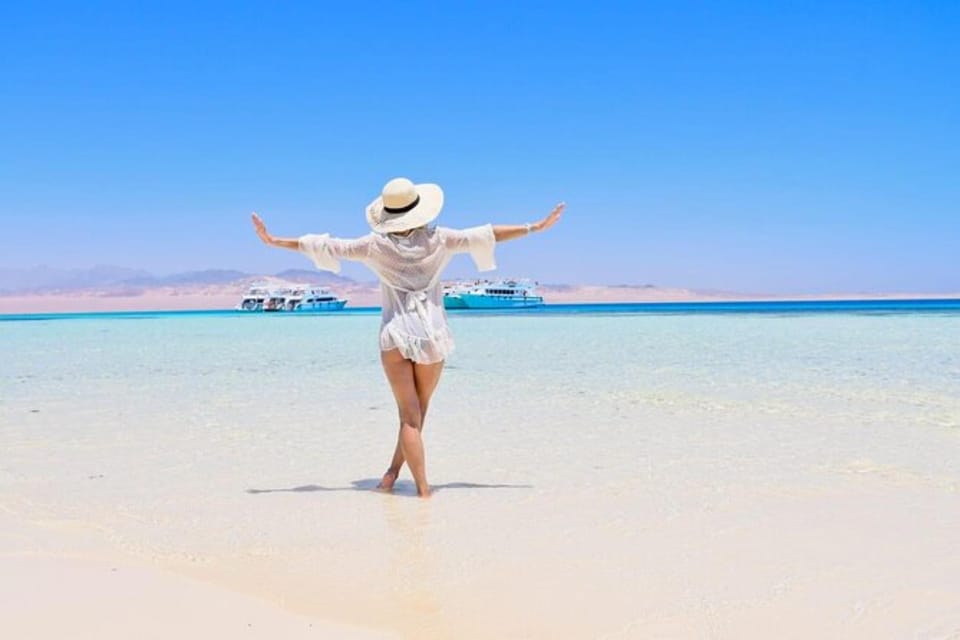 A woman standing ankle-deep in glass-clear shallow water on a pale sandbank, arms held wide, with moored excursion boats and low desert mountains in the distance