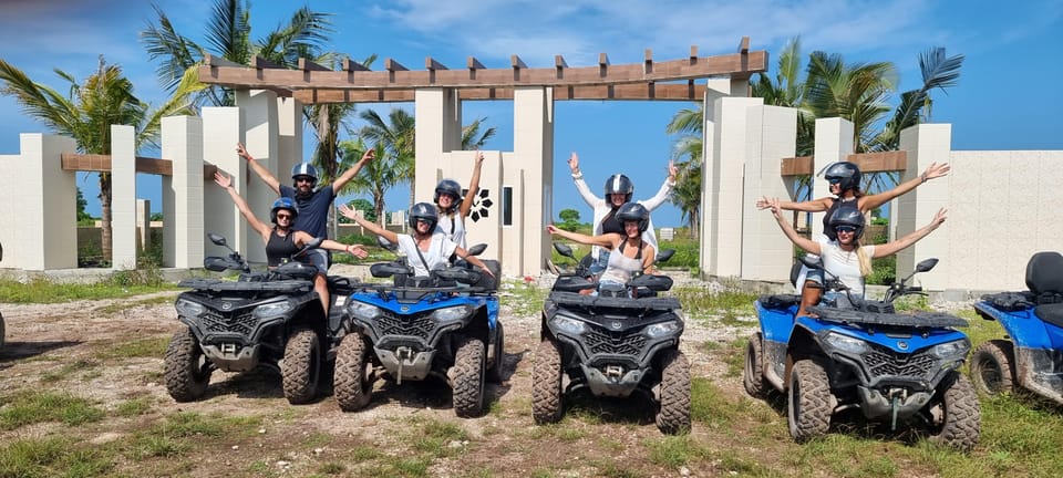 Riders in helmets cheering with raised arms from four parked quad bikes in front of a pale stone gateway