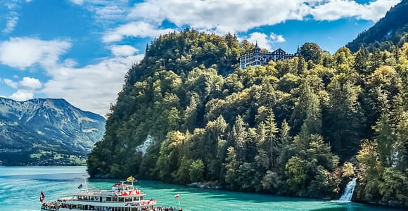 A white passenger boat on turquoise water below a forested slope with a hotel on top and a small waterfall
