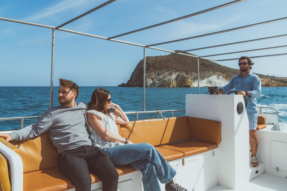 Two passengers on a covered boat deck, the skipper at the wheel, with a pale headland rising from the sea behind