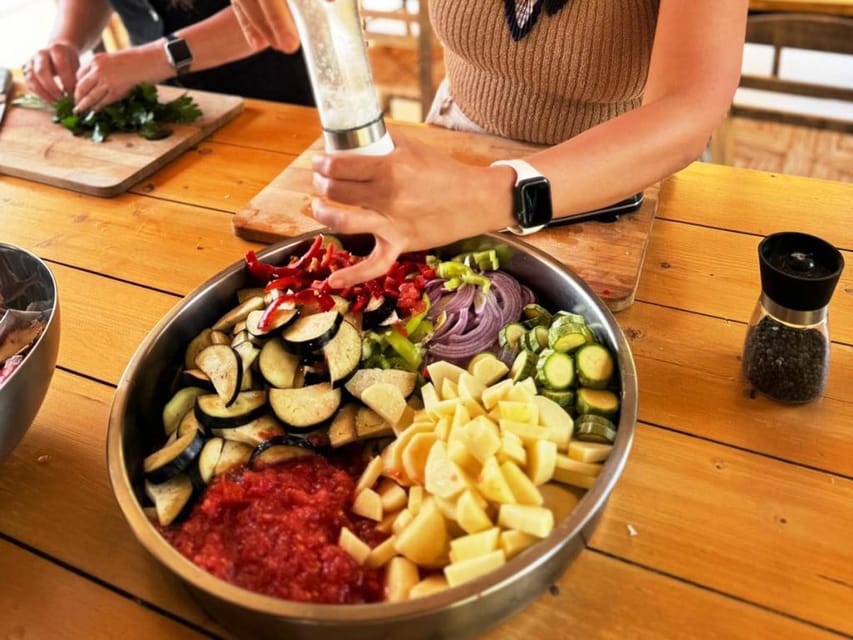 Sliced aubergine, potato, courgette, red pepper and onion in a steel bowl on a wooden table, with a hand grinding pepper over it