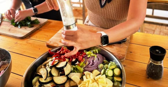 Sliced aubergine, potato, courgette, red pepper and onion in a steel bowl on a wooden table, with a hand grinding pepper over it