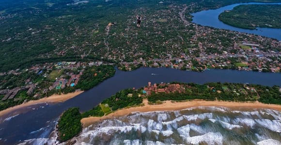 Aerial view of a river meeting the sea beside a long sandspit and beach, a town of red roofs and palms inland, a paramotor canopy in the sky