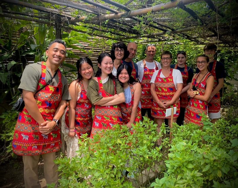 A dozen course participants in red elephant-patterned aprons standing together under a bamboo pergola in a herb garden