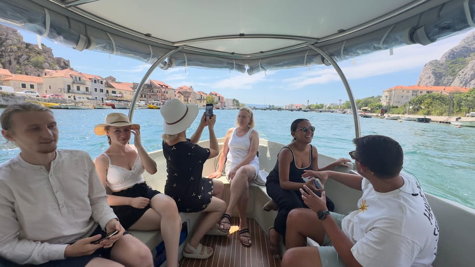 Passengers sit under a canopy on a boat, with the houses of a coastal town behind them