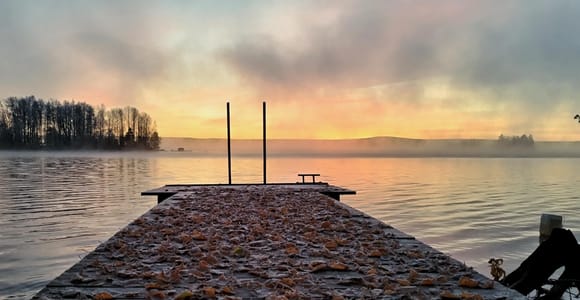 A wooden sauna cabin with steam rising beside a snowy riverbank at dusk