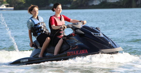 Two people in life jackets riding a jet ski across flat lagoon water, palm trees and hotel roofs along the far bank