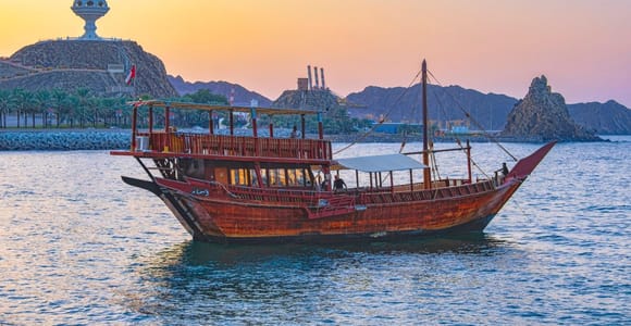 A wooden dhow with a white awning lies at anchor in calm water at dusk, the ornamental incense-burner monument on a rocky headland behind it and orange sky over dark mountains