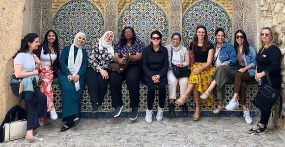 A group of women sitting side by side in front of a tiled niche with a green-tiled roof, set into an old stone wall