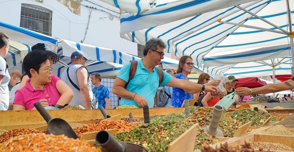 Visitors leaning over wooden bins of dried herbs and spices at a market stall under blue and white awnings