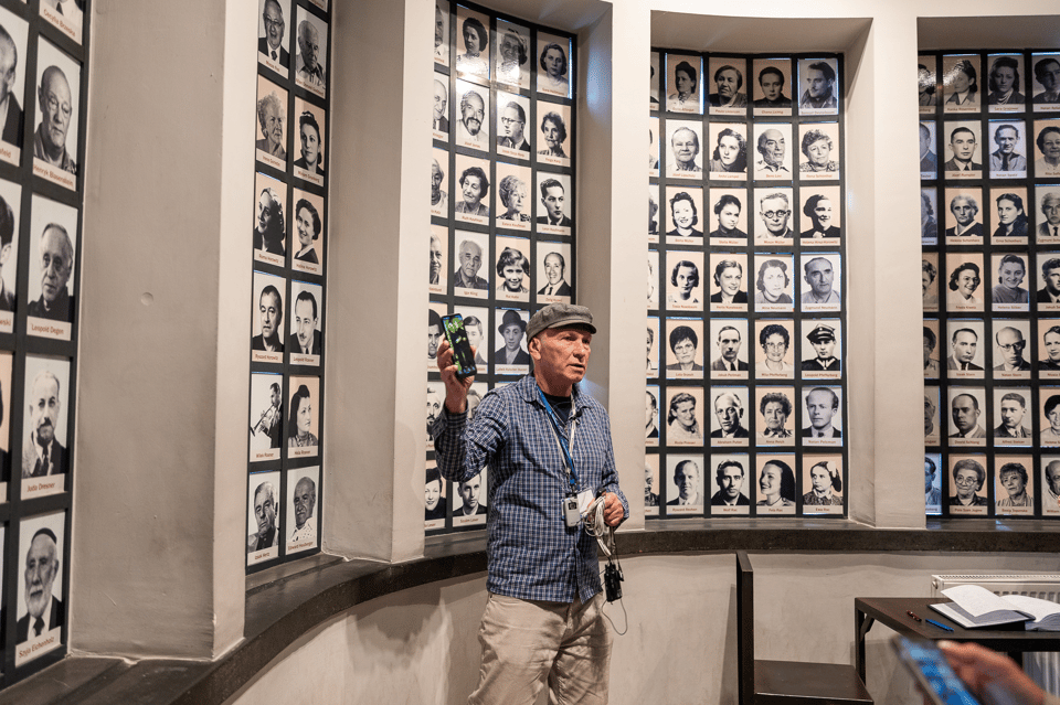 A guide in a cap holds up a phone in front of walls of framed black-and-white portrait photographs