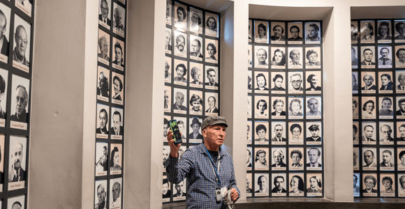 A guide in a cap holds up a phone in front of walls of framed black-and-white portrait photographs