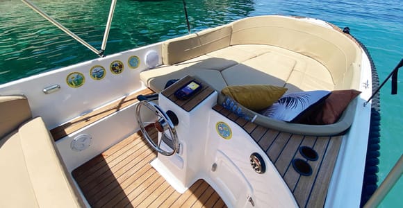 Open motorboat seen from the helm, a steering wheel and cream cushioned seats, turquoise water beside the hull