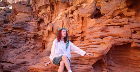 A woman in a white shirt and headscarf sitting on a ledge of layered orange and red sandstone, the rock wall curving above and around her