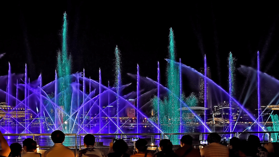 Jets of water lit purple and green rising over the bay during an evening show, seen over the heads of an audience standing at the waterfront railing
