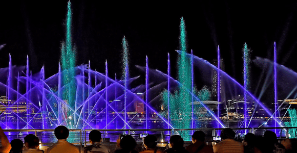 Jets of water lit purple and green rising over the bay during an evening show, seen over the heads of an audience standing at the waterfront railing