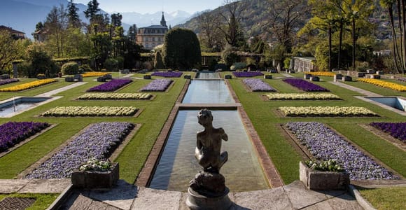 Formal terraced garden with a long water channel, a bronze statue, lawns and flower beds in purple and yellow, a villa among trees and snowy mountains behind
