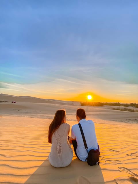 Two people sitting on a sand ridge with their backs to the camera, watching the sun sink behind low dunes