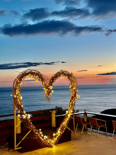 A lit heart made of twigs and flowers stands on a terrace above the sea at sunset