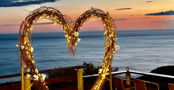 A lit heart made of twigs and flowers stands on a terrace above the sea at sunset