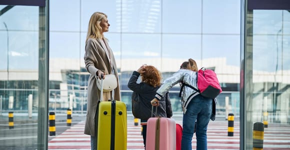 A woman and two children with suitcases and a backpack stand at a glass wall and look out at a crossing in front of an airport terminal