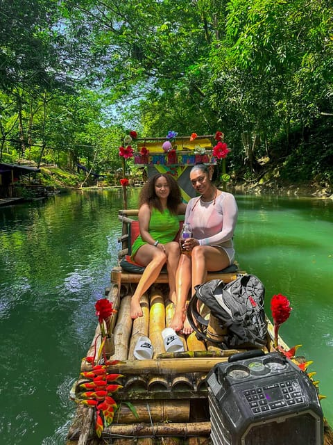 Two passengers sitting on a flower-decorated bamboo raft drifting down a narrow green river between overhanging trees