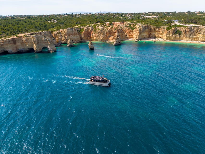 White catamaran carrying passengers beside layered golden cliffs above deep blue water