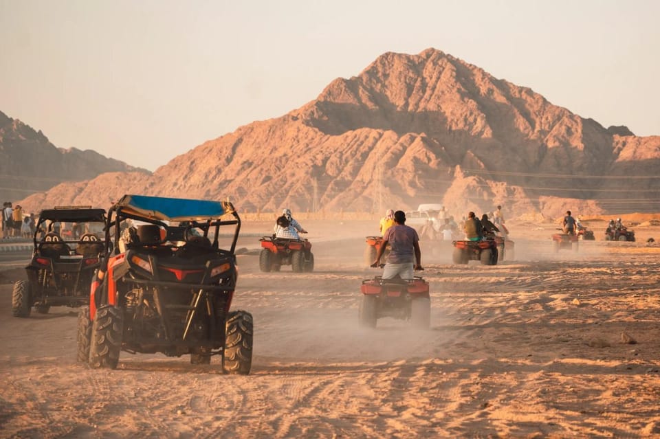 Una fila di quad e un buggy arancione che sollevano polvere su una pista nel deserto, con una cresta montuosa brulla e marrone sullo sfondo nella luce bassa della sera