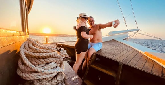A couple taking a selfie on the wooden deck of a sailing boat, with the low sun setting over a calm sea behind them