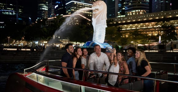 The open deck of a night cruise boat on the river, the Merlion spouting into the water behind and the lit towers of the financial district above