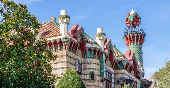 A villa with red brick, tiled green and red bands and a round tower topped with a tiled cap, seen through trees under a blue sky