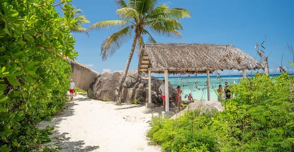 A sandy path between low green bushes leading past granite boulders to a thatched shelter and pale turquoise water