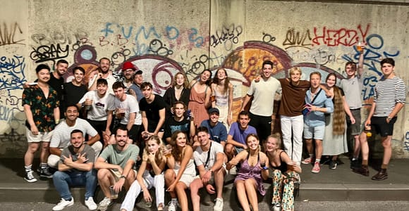 A large group posing at night on a pavement in front of a wall covered in graffiti