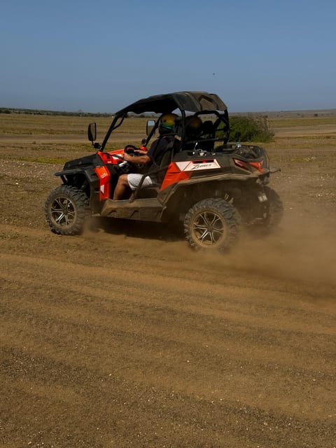 An orange and black two-seater buggy throwing up a long plume of dust on a flat dirt track under a clear sky