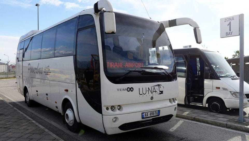 Biały autokar z tabliczką lotniska w Tiranie za szybą, zaparkowany na stanowisku obok mniejszego busa