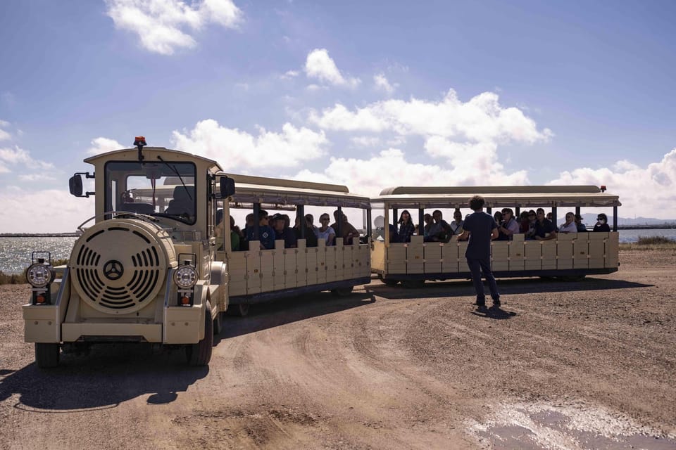A small tourist train with open carriages full of passengers and a person standing beside it on a dirt track by the lagoon