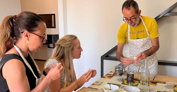 Two women in aprons shaping filled pasta by hand while a man in a yellow shirt demonstrates beside a pasta machine on a table