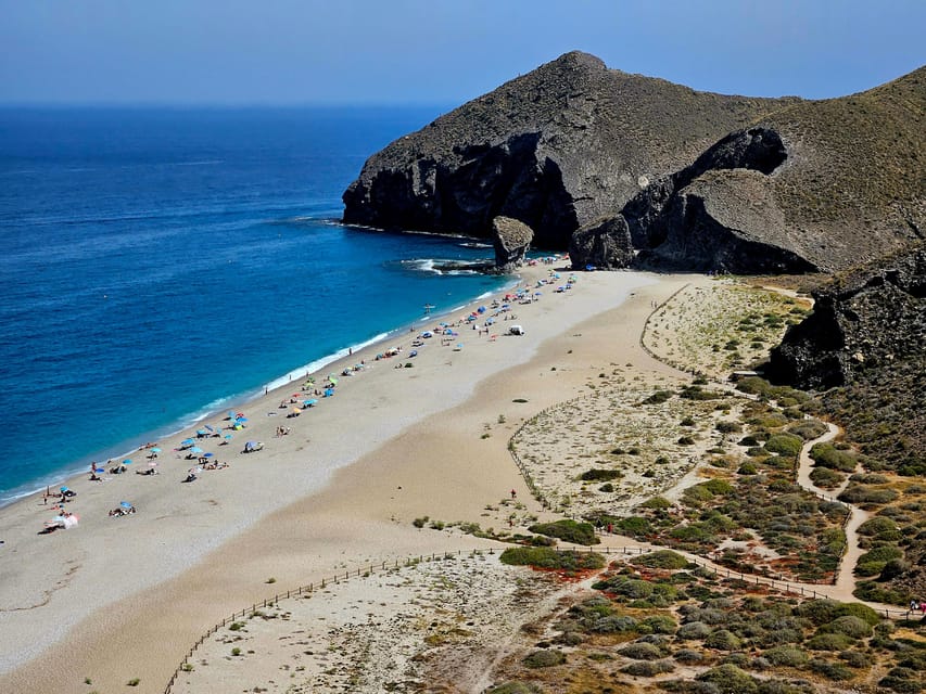 A pebbly cove at the foot of a tall cliff, with clear water and few people