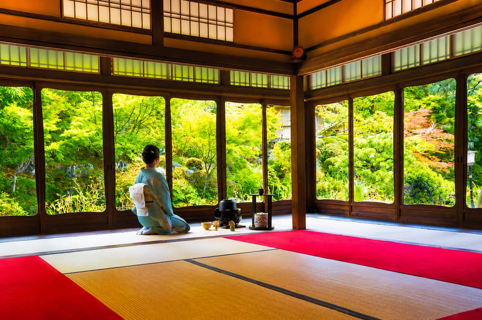 A woman in a pale blue kimono kneeling on tatami before a small hearth, facing a wall of open windows onto a green maple garden